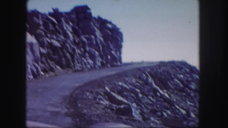 Colorado mountain road in a 1958 8mm frame, with a rocky cliff face, curved pavement, and soft motion blur from the camera.