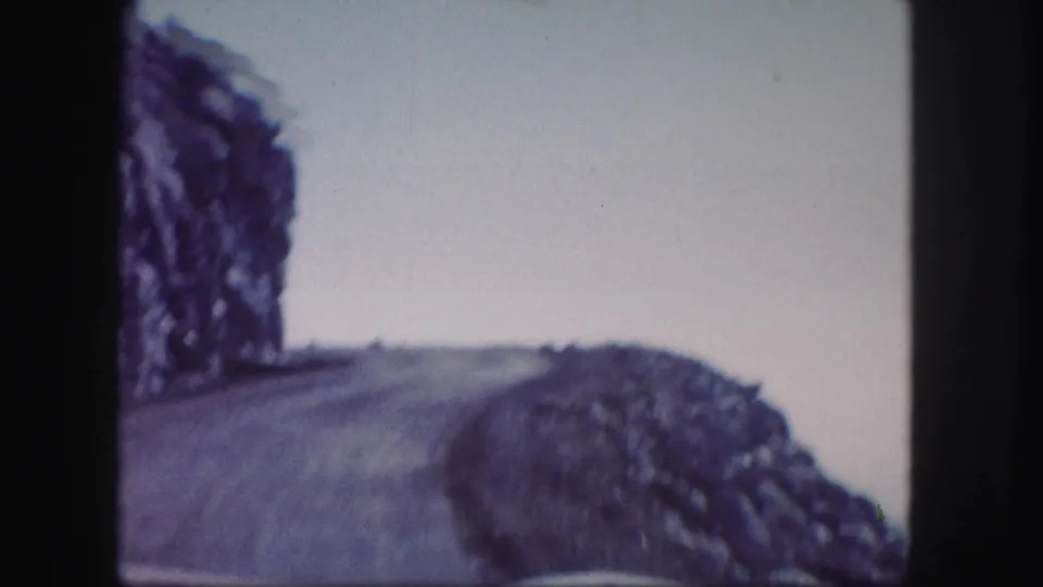 1958 Colorado 8mm frame showing a curving mountain road bordered by rocky terrain beneath an overcast sky.
