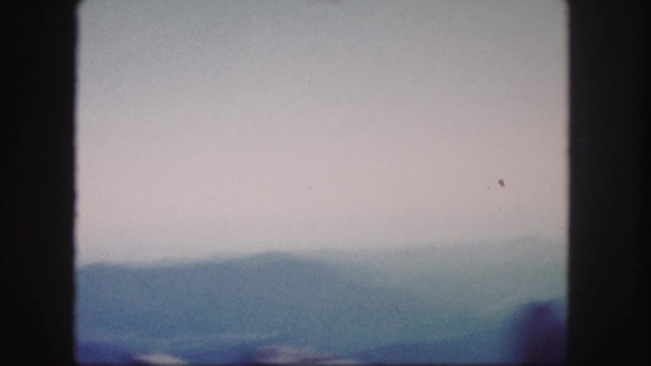 1958 Colorado home-movie frame with layered distant mountains, pale sky, and a blurred dark ridge near the lower right.