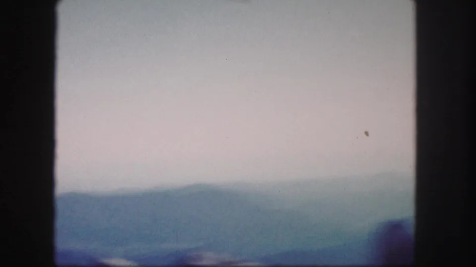 1958 Colorado home-movie frame with layered distant mountains, pale sky, and a blurred dark ridge near the lower right.