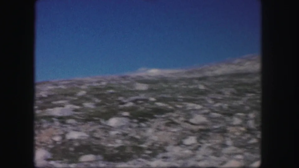 1958 Colorado 8mm frame showing a stony, treeless hillside with sparse low vegetation beneath a clear blue sky.
