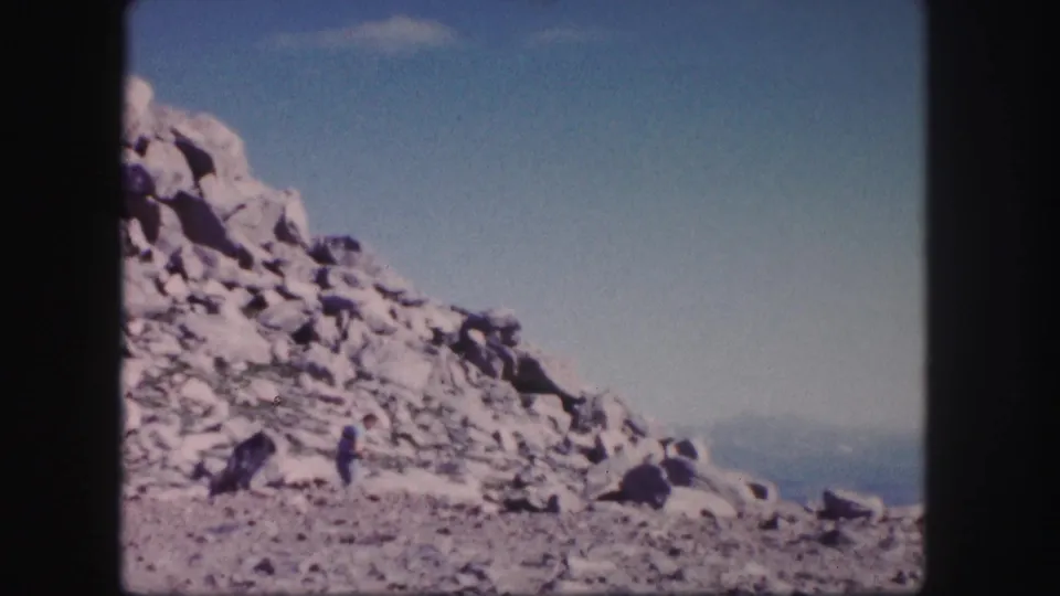 1958 Colorado 8mm frame of a craggy mountain slope with a lone distant hiker standing among rocks beneath blue sky.