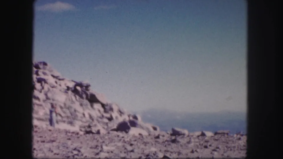 1958 Colorado 8mm frame of a rocky hillside with a distant hiker partly near the left edge and hazy mountains beyond.