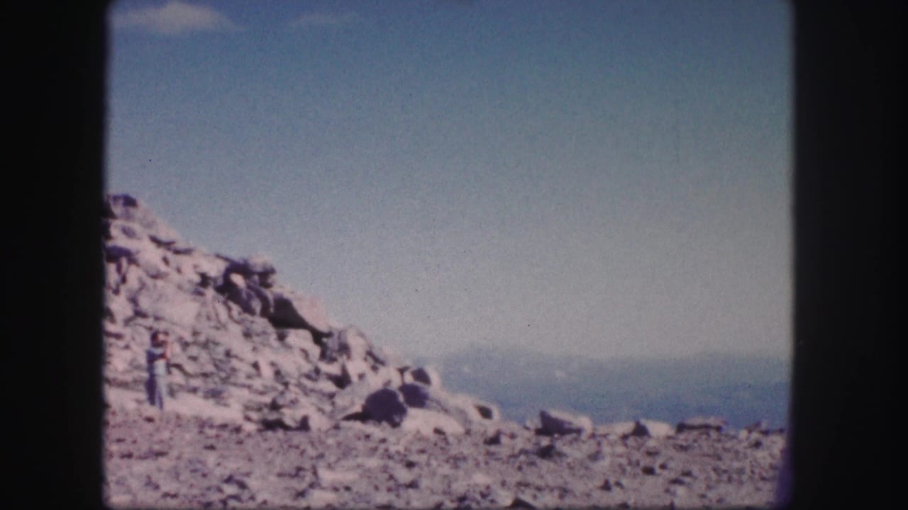 1958 Colorado 8mm frame showing a rocky slope, hazy distant peaks, and a person standing near the left side of the rocks.