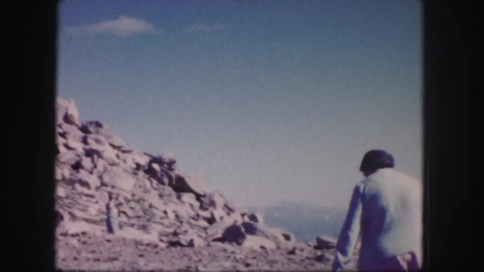 1958 Colorado 8mm frame showing a person walking across the foreground, with rocks, sky, and distant peaks behind.