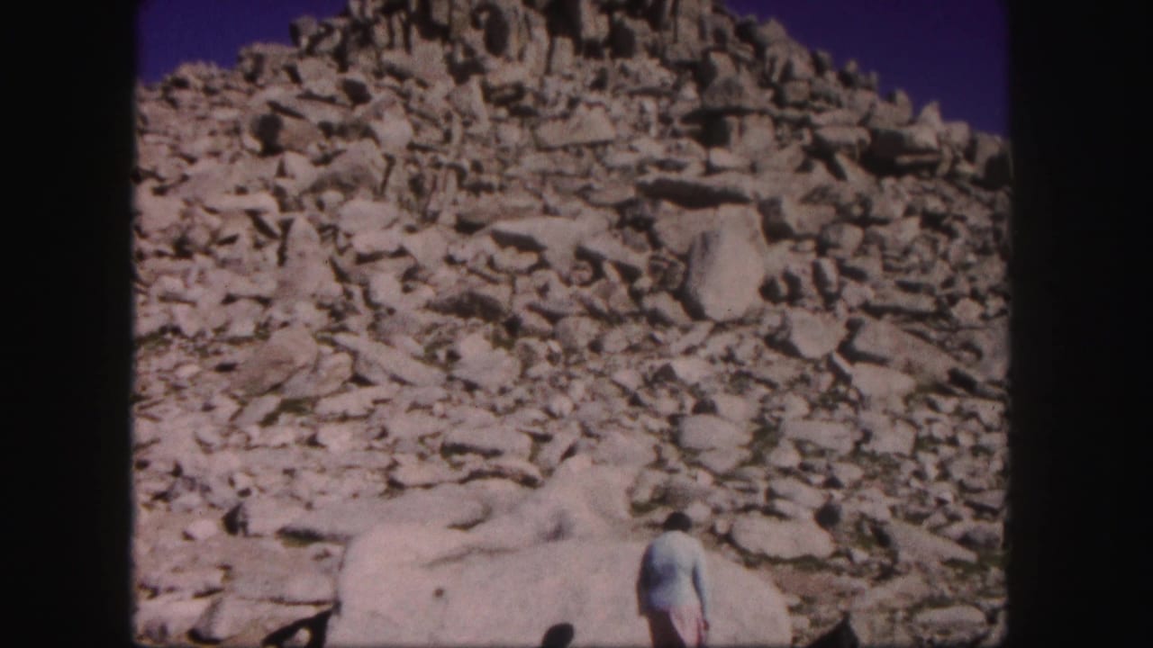 1958 Colorado 8mm frame of a woman in a light top near pale boulders beneath a rugged scree-covered mountain slope.