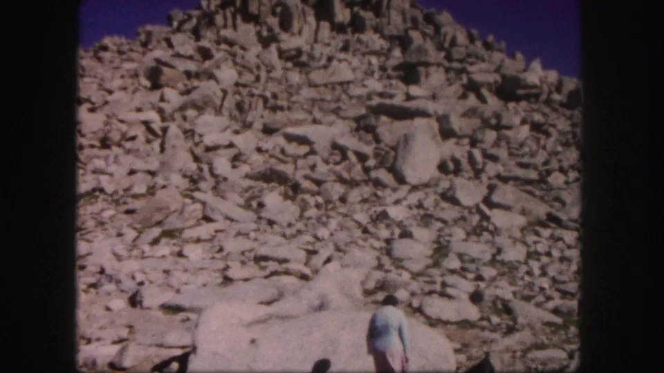1958 Colorado 8mm frame of a woman in a light top near pale boulders beneath a rugged scree-covered mountain slope.