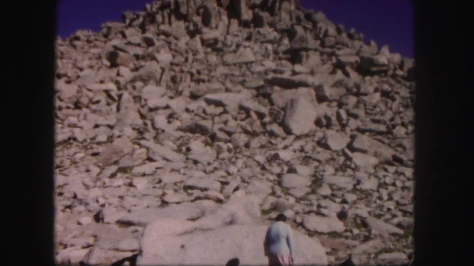 1958 Colorado home-movie frame showing a woman at the lower edge of a boulder field below a rugged mountain peak.
