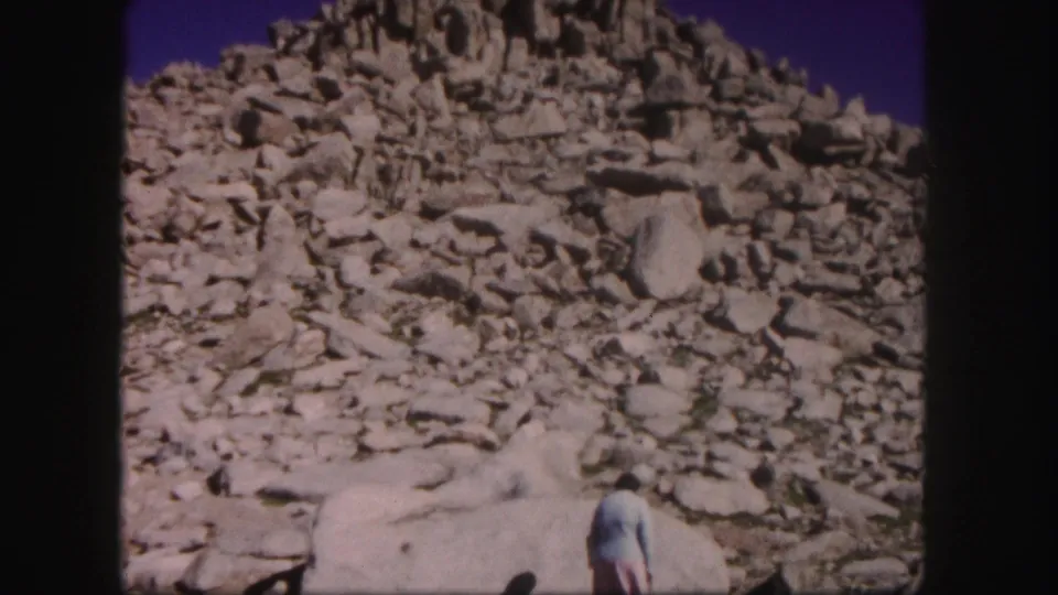 1958 Colorado home-movie frame of a woman at lower right below sunlit boulders and a jagged scree-covered slope.