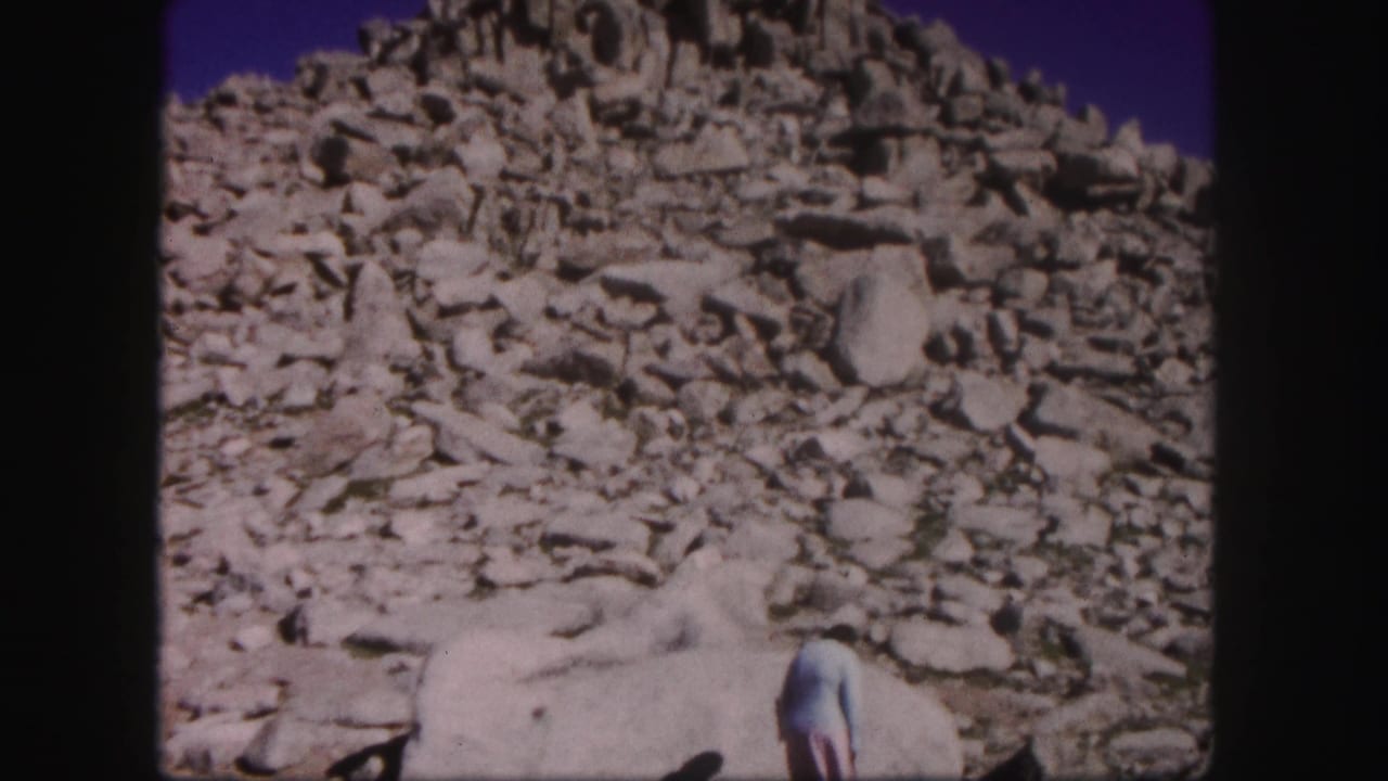 1958 Colorado home-movie frame with a rocky summit slope and a person partly cropped at the bottom of the frame.