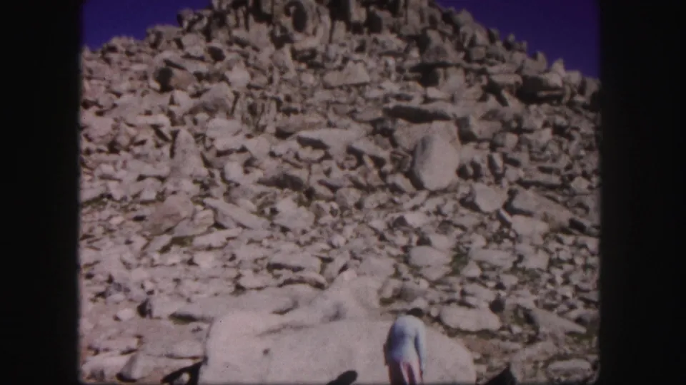 1958 Colorado home-movie frame with a rocky summit slope and a person partly cropped at the bottom of the frame.