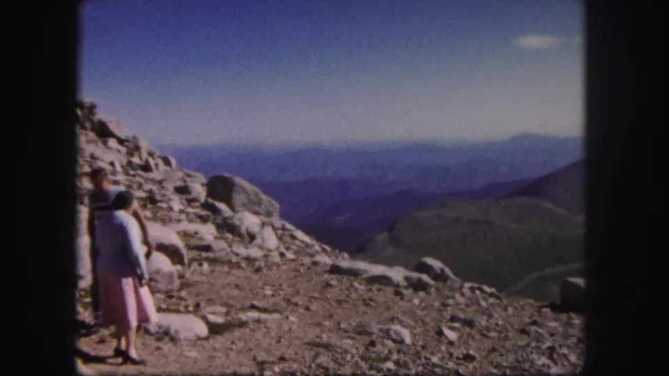 1958 Colorado home-movie frame with a couple near loose rocks, facing a broad hazy mountain landscape under blue sky.