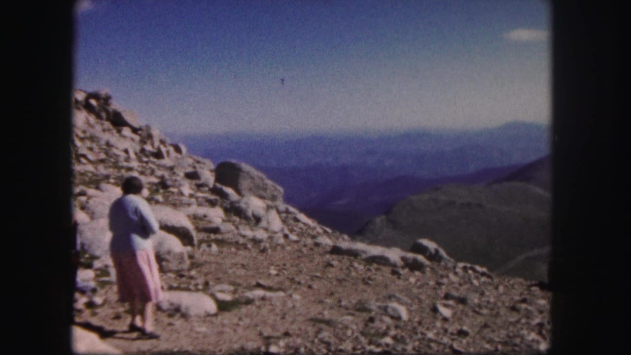 1958 Colorado 8mm frame showing a woman in a light sweater and pink skirt on rocky summit terrain with layered mountain views.