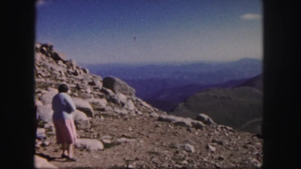 1958 Colorado 8mm frame showing a woman in a light sweater and pink skirt on rocky summit terrain with layered mountain views.