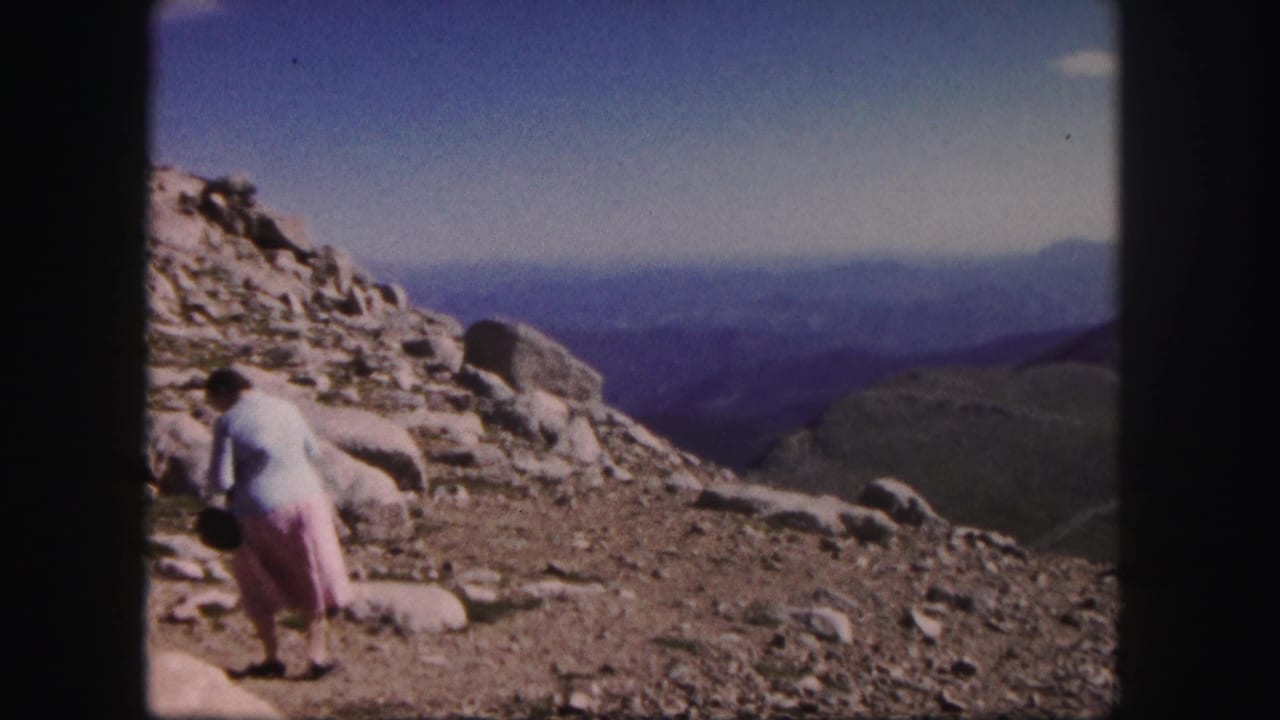 Woman bending near rocks on a mountain slope with broad ridges in the background.
