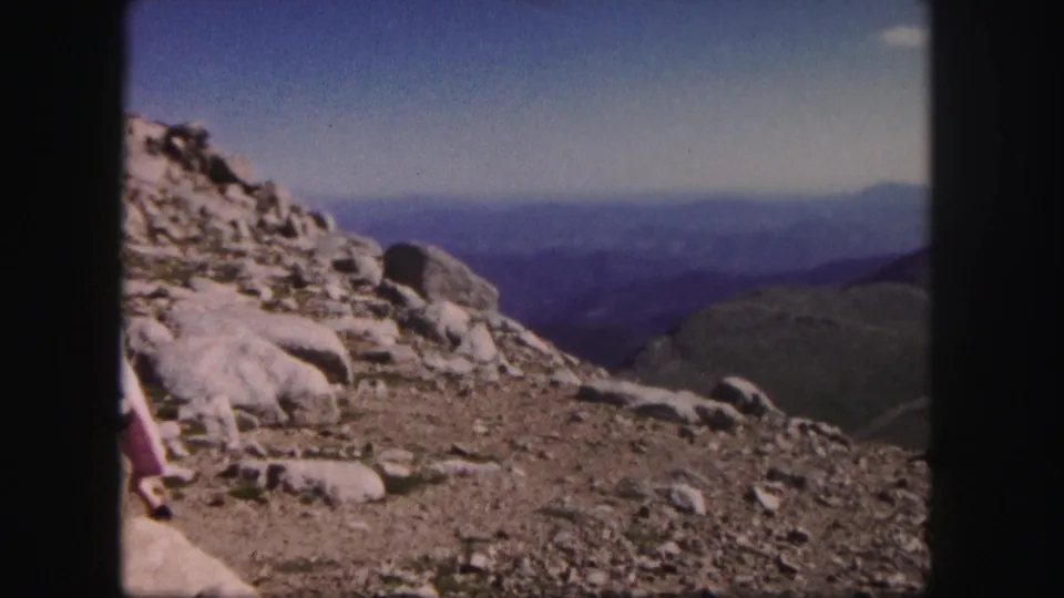 Rocky Colorado summit terrain fills the frame, with the woman partly exiting at left and layered mountain ridges in the distance.