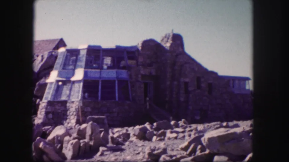 Home-movie frame from 1958 Colorado showing the building closer, with stone walls, glass windows, and boulders below.