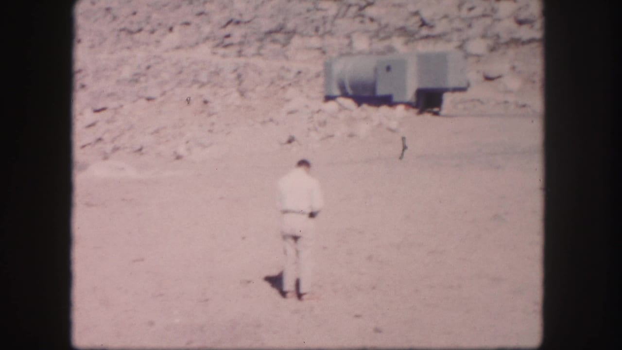 Man in a light suit stands on open ground with a trailer and rocky hillside behind him.