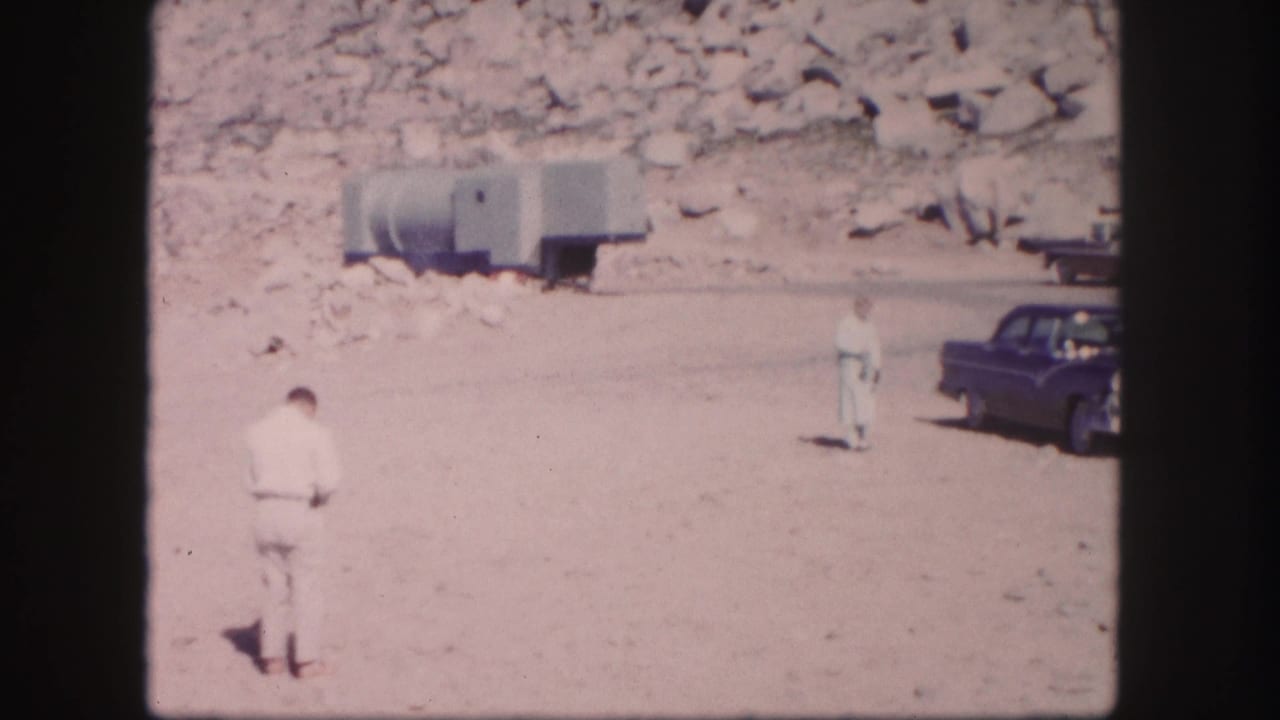 1958 Colorado home-movie frame with a man in a light suit, a second man near the road, a dark sedan, and a trailer by the rocks.