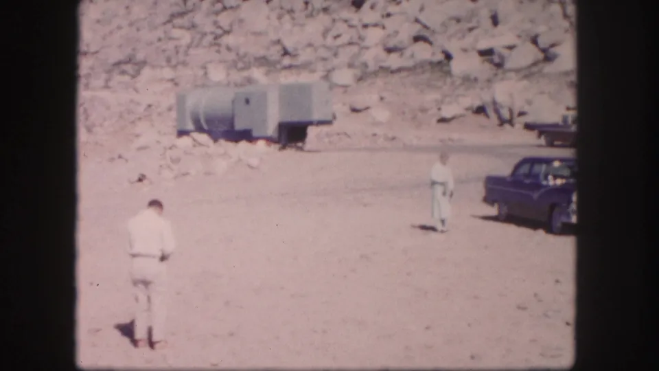 1958 Colorado home-movie frame with a man in a light suit, a second man near the road, a dark sedan, and a trailer by the rocks.
