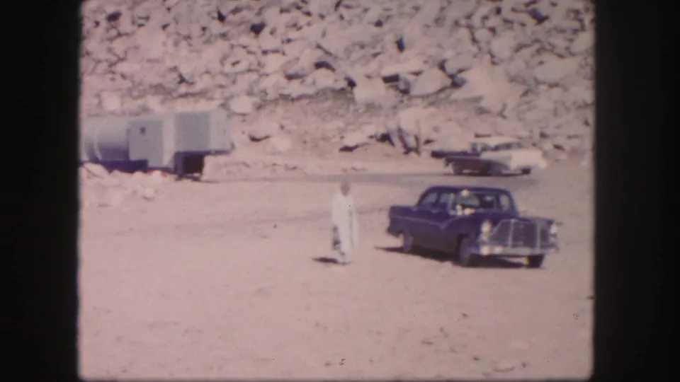 1958 Colorado frame showing a blue classic sedan beside a man in pale clothing, with another vehicle, trailer, and rocky hillside beyond.