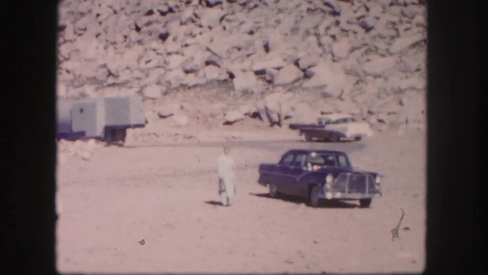 1958 Colorado 8mm frame with a blue 1950s sedan, a man in light clothing, a trailer, and another vehicle against a rocky hillside.