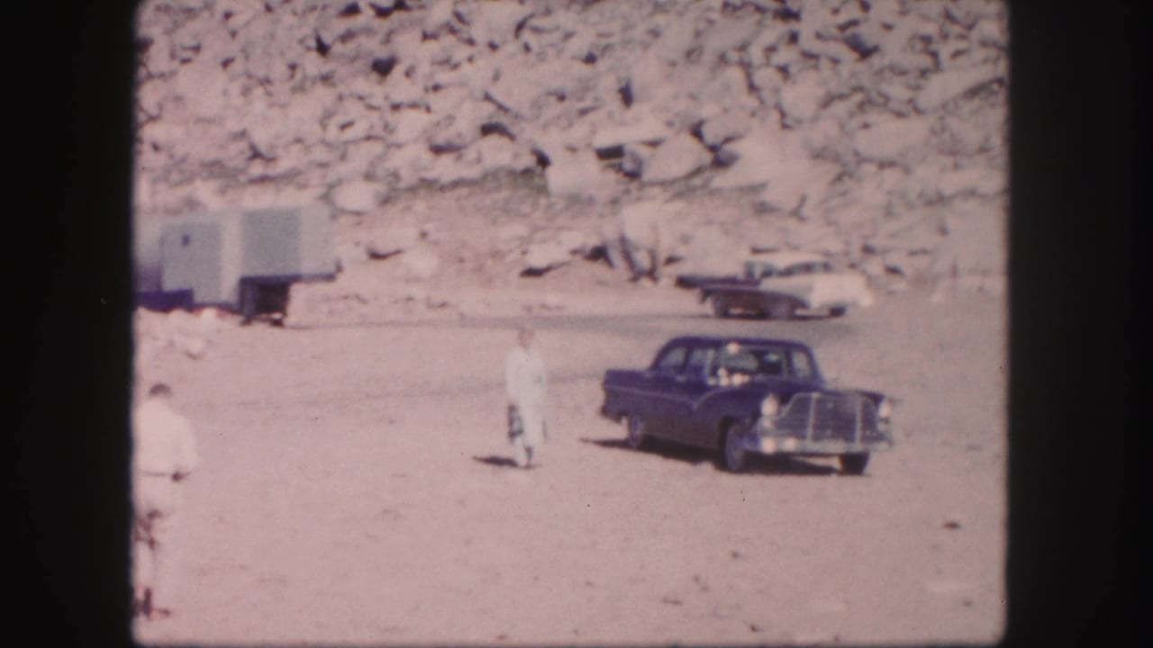 1958 Colorado home-movie frame showing a dark car near a rocky mountain roadside with a person standing in bright sun.