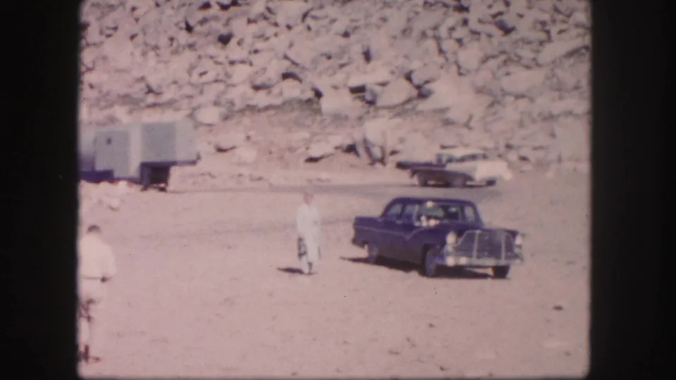 1958 Colorado home-movie frame showing a dark car near a rocky mountain roadside with a person standing in bright sun.