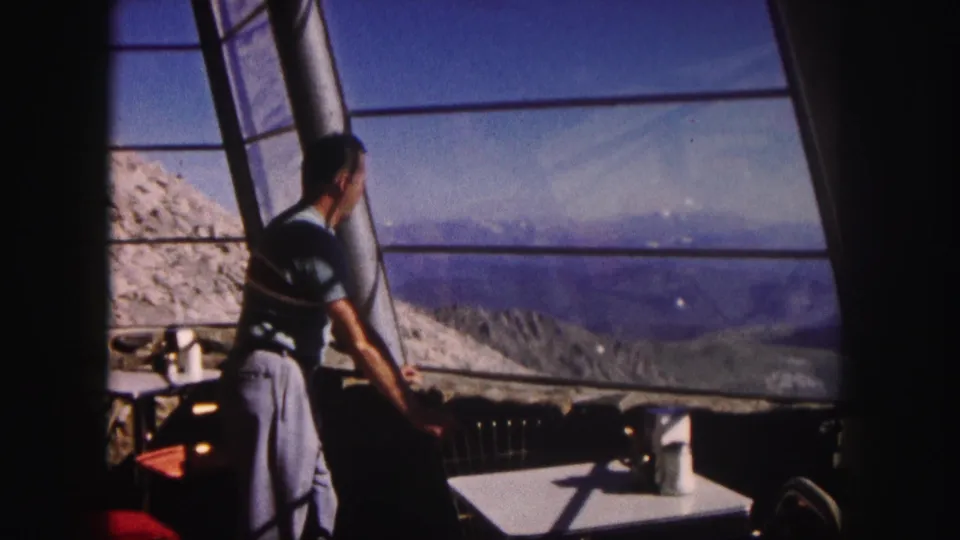 1958 Colorado frame of a man in a striped shirt inside a mountain-view room, leaning near a table and looking through large windows.