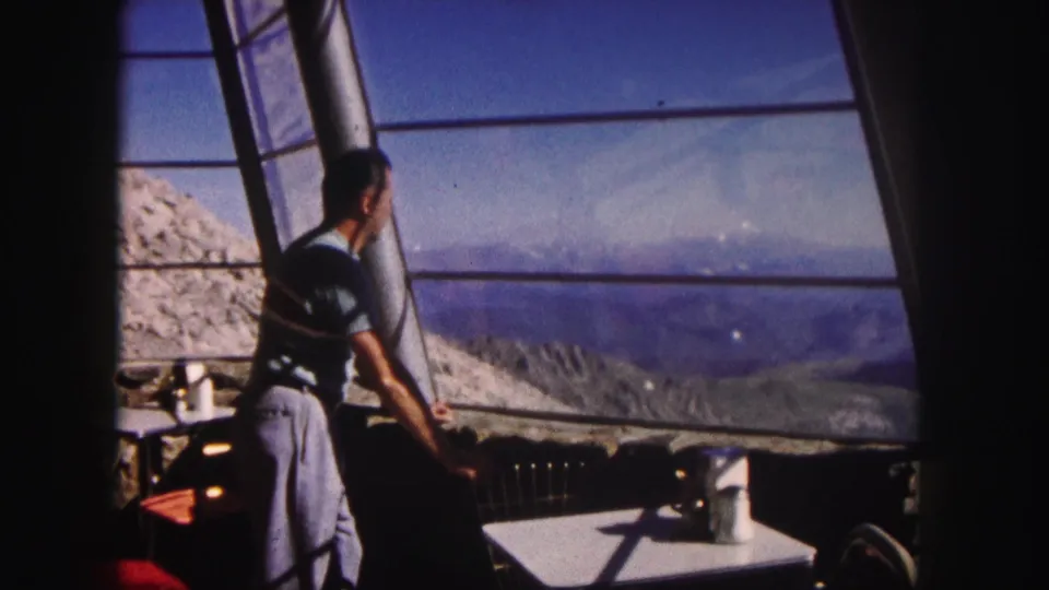1958 Colorado home-movie frame showing a man beside broad windows, with interior seating and a rugged mountain panorama outside.