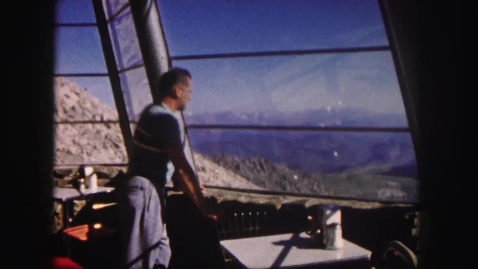 1958 Colorado frame of a man leaning near a table or counter inside a sunlit room with mountains visible through the windows.
