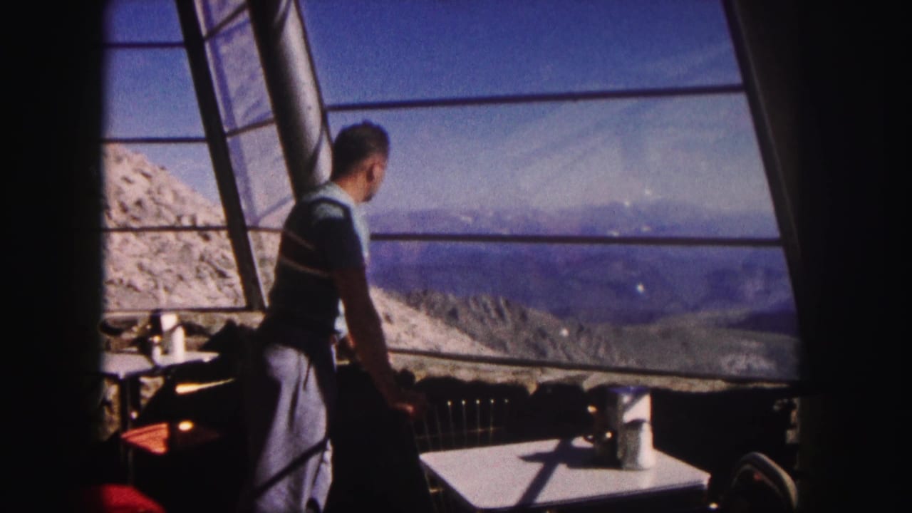 1958 Colorado home-movie frame of a man in a striped shirt looking through glass panels toward a mountain landscape.