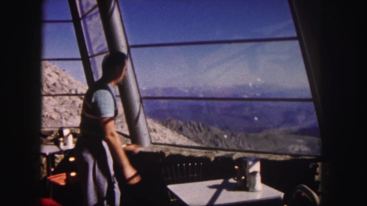 Man stands near a large window inside a viewpoint with mountains beyond the glass.