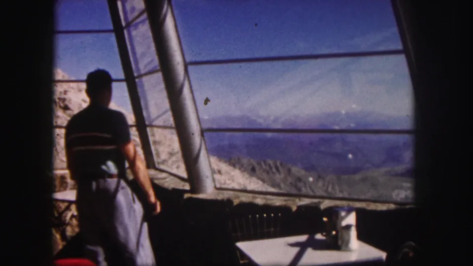 From behind, a man watches the Colorado mountain landscape through the angled glass and metal framing of a viewpoint.