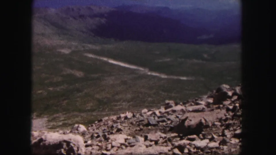 Colorado mountain vista on 8mm film, with rocky foreground, distant slopes, and a pale road winding through the landscape.