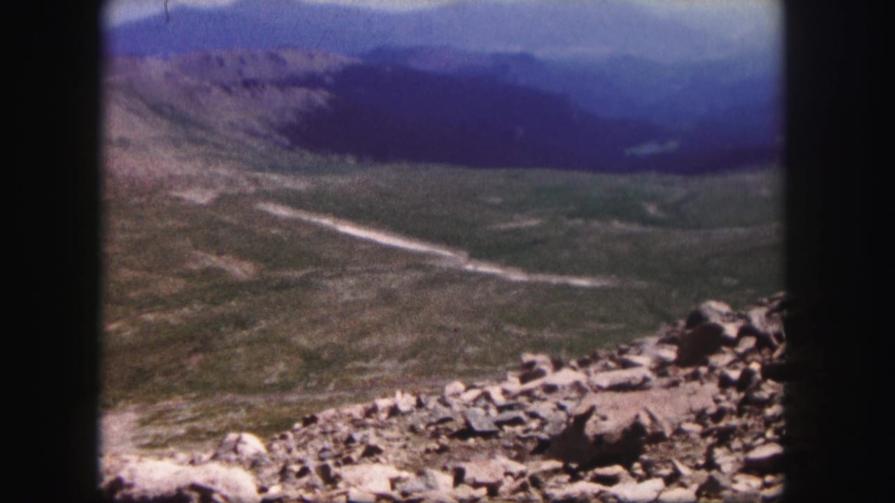 Mountain valley view with rocky foreground and a light road crossing the terrain.
