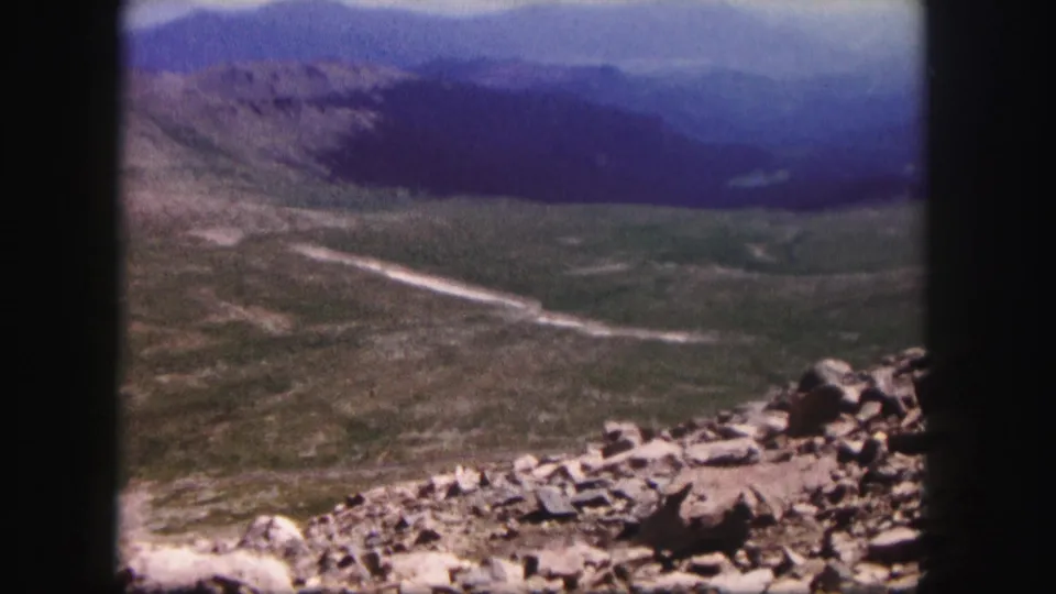 1958 Colorado 8mm frame with visible grain, rocky ridge foreground, broad valley, winding road, and distant mountains.