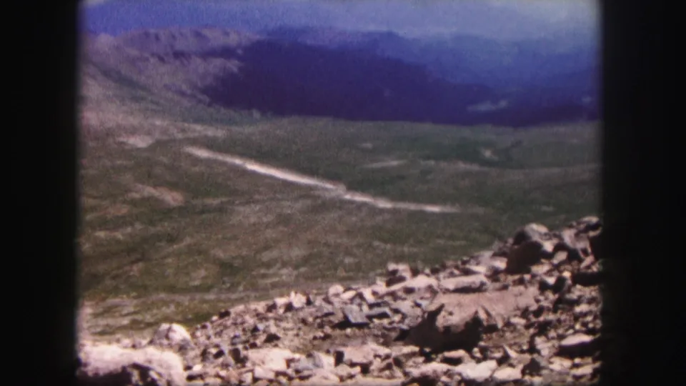 1958 Colorado frame showing a steady home-movie view over rocks, an open valley, a winding dirt road, and distant hills.