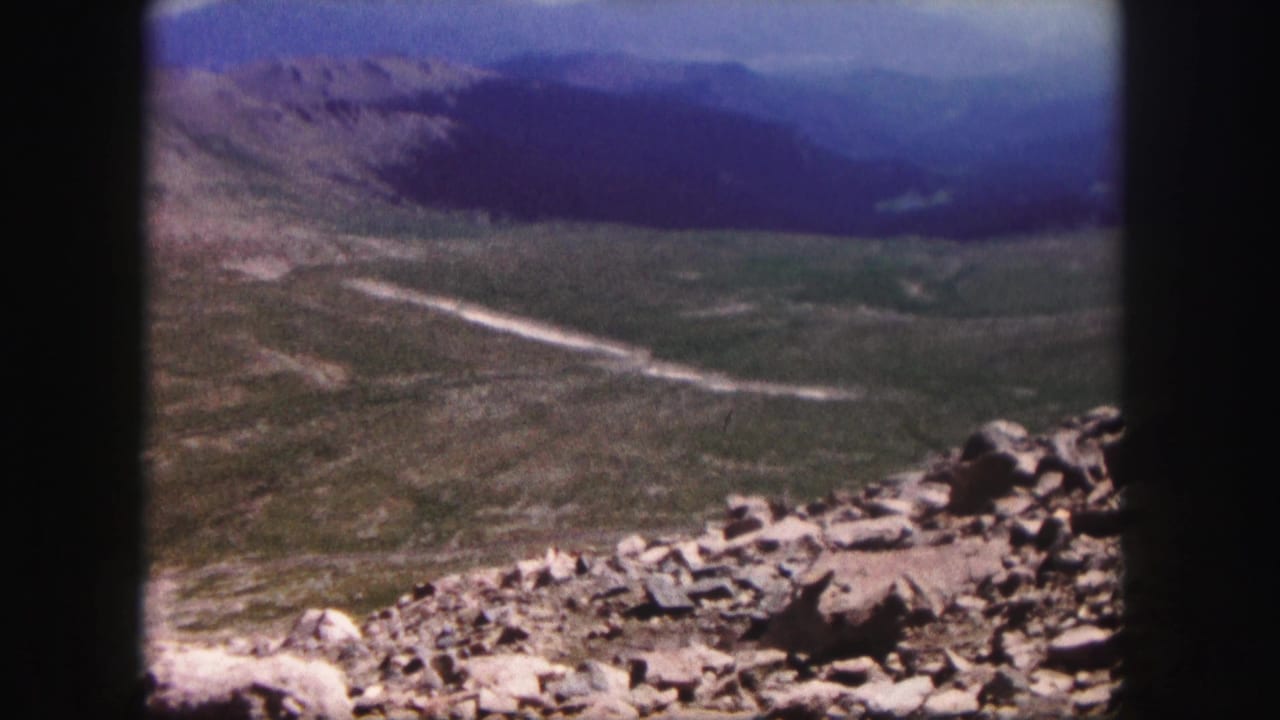 1958 Colorado 8mm frame showing a rocky summit foreground above a broad mountain valley and distant hazy ridgelines.