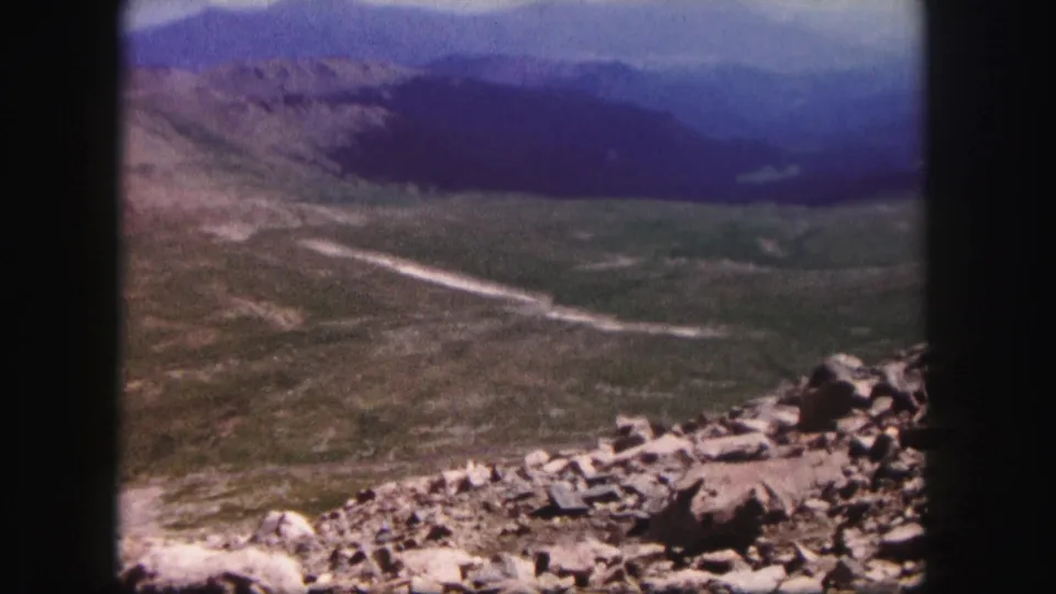 1958 Colorado home-movie frame of a rocky overlook facing a broad valley, distant mountains, and a pale track or stream below.