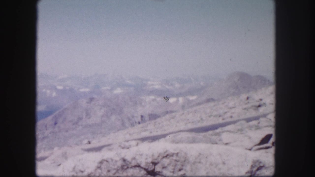1958 Colorado home-movie frame showing a snow-streaked mountain ridge, rocky foreground, and layered peaks fading into haze.