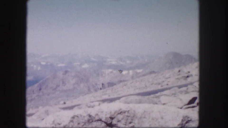 1958 Colorado home-movie frame showing a snow-streaked mountain ridge, rocky foreground, and layered peaks fading into haze.
