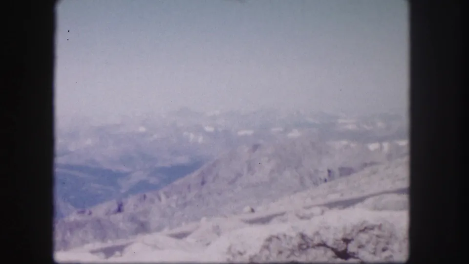 Vintage 8mm Colorado frame showing a hazy mountain panorama, snow-streaked foreground slope, and broad pale sky.