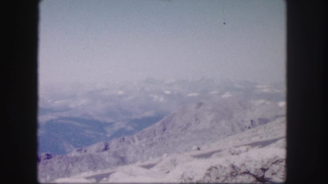 1958 Colorado 8mm frame showing distant hazy mountains beyond a rocky, snow-dusted ridge under a pale blue sky.