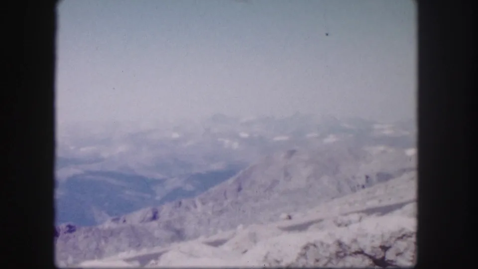 1958 Colorado 8mm frame showing distant hazy mountains beyond a rocky, snow-dusted ridge under a pale blue sky.