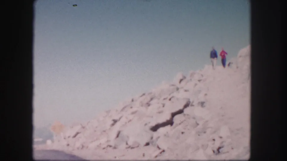 Two distant hikers in blue and red outerwear are visible on a rocky Colorado ridge in the final 1958 home-movie frame.