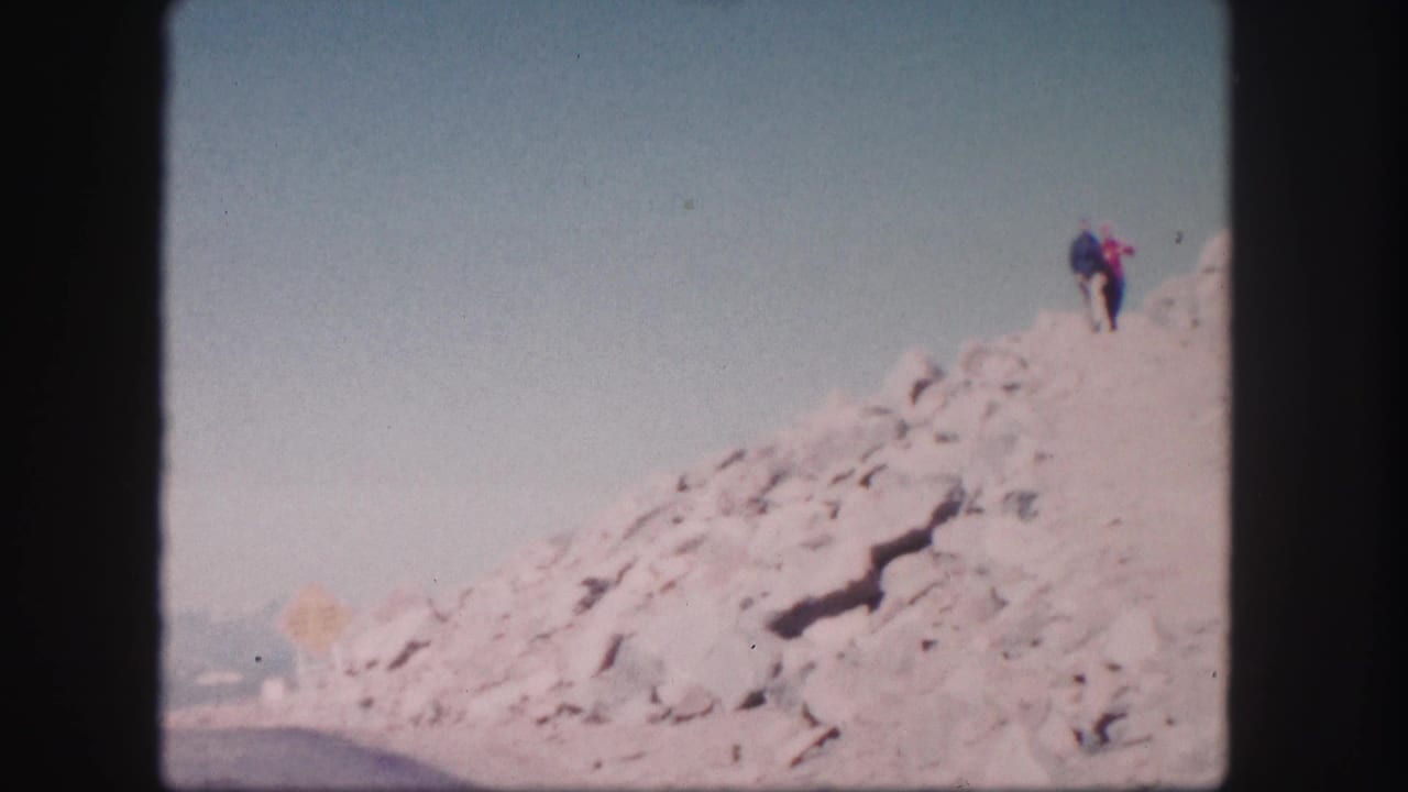 Two distant people stand on a pale rocky slope above a road under a clear sky.