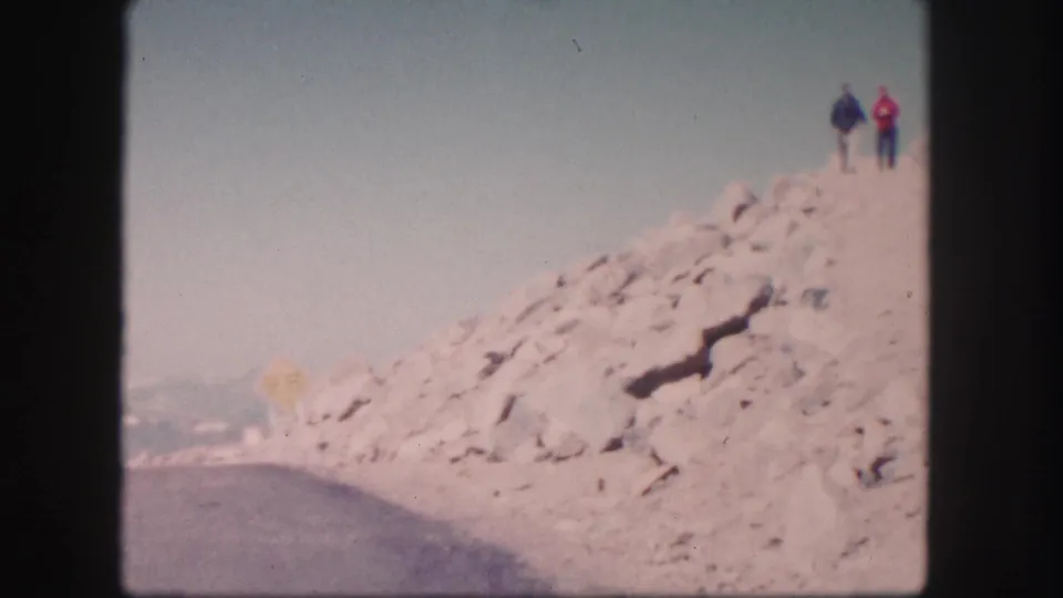 Two people walk along a rough rocky slope above a curving road in Colorado, captured on faded 1958 8mm film.
