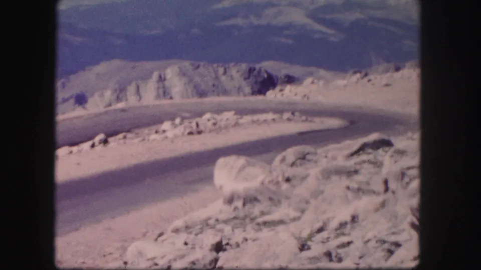 A winding Colorado mountain road curves through pale rocky terrain, with distant ridges visible in 1958 8mm home-movie footage.