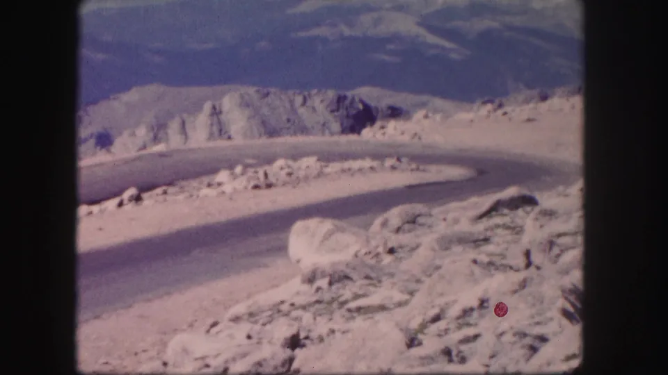 1958 Colorado 8mm frame showing a road bend beside boulders and pale rugged terrain, with blue sky above.